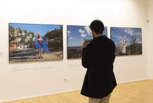 Una sala de exposición de arte en ARCO Madrid una persona observando y fotografiando diversas obras de arte en las paredes. La gente conversa y admira las impresiones artísticas y fotográficas.