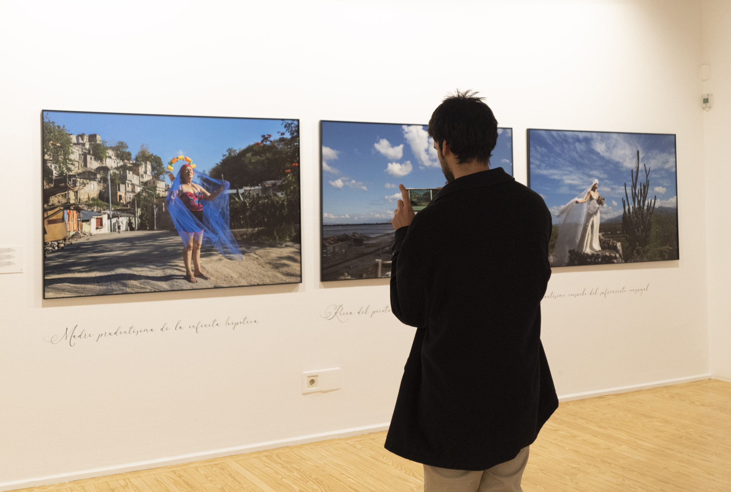 Una sala de exposición de arte en ARCO Madrid una persona observando y fotografiando diversas obras de arte en las paredes. La gente conversa y admira las impresiones artísticas y fotográficas.