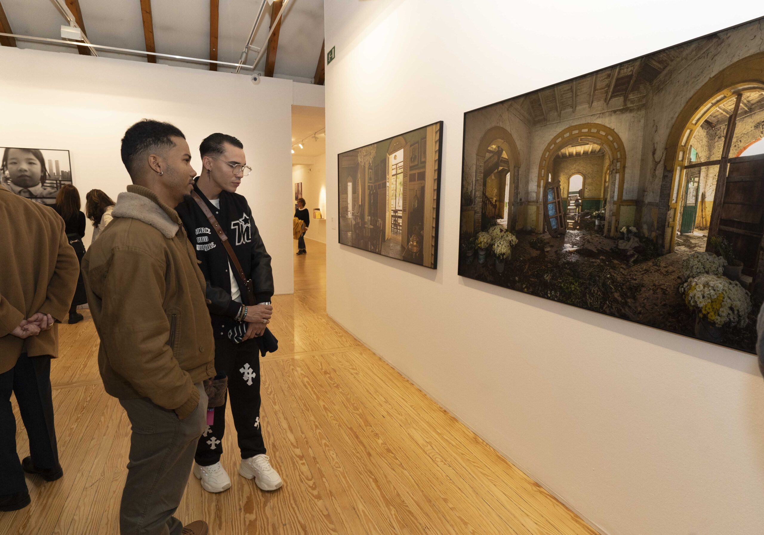 Una sala de exposición de arte en ARCO Madrid dos personas observando diversas obras de arte en las paredes. La gente conversa y admira las impresiones artísticas y fotográficas.